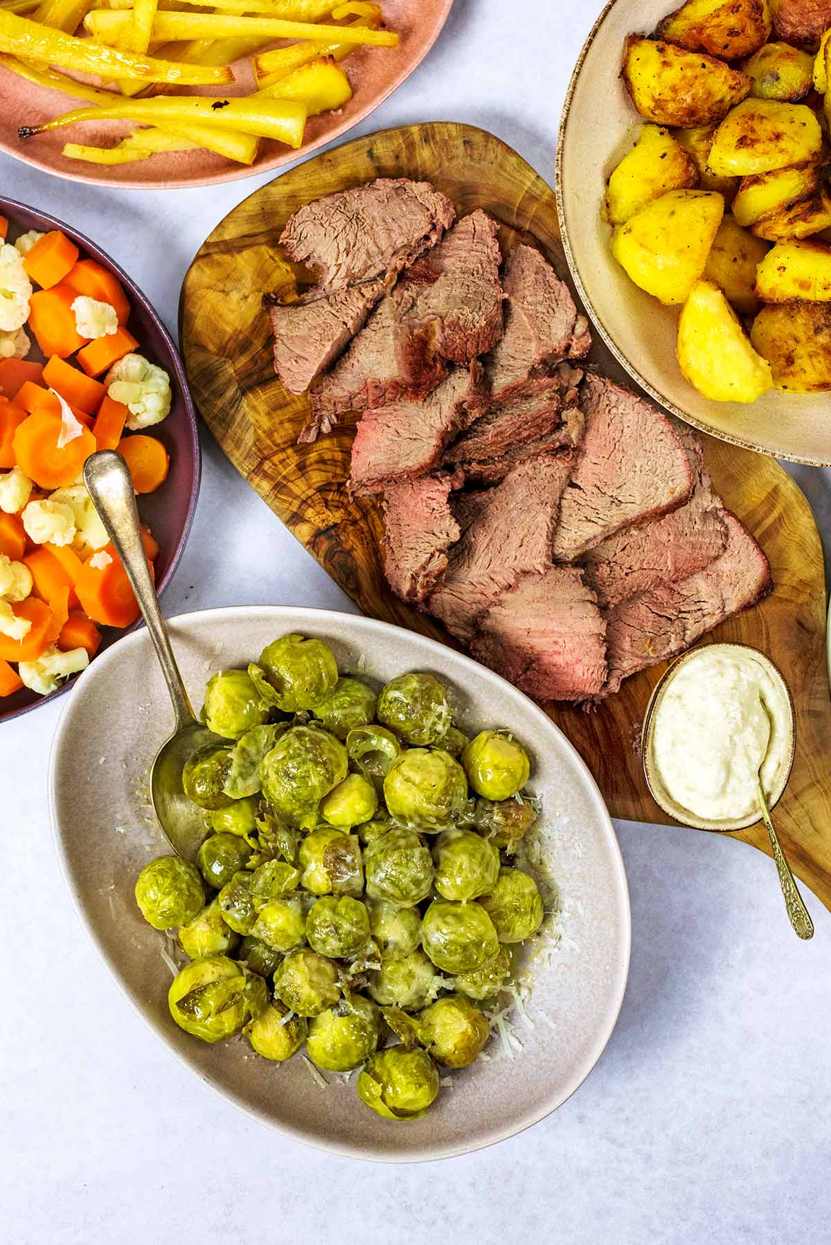 A bowl of Brussels sprouts next to a board of roast beef, some potatoes, carrots and parsnips.