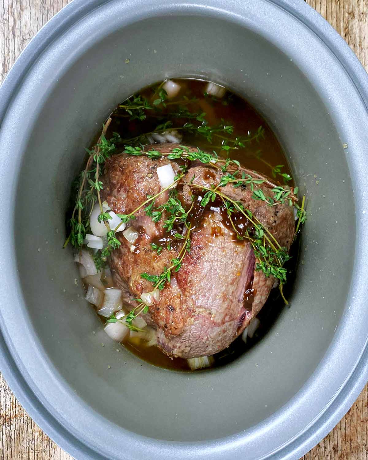 Browned beef joint in a slow cooker with thyme, shallots and stock.