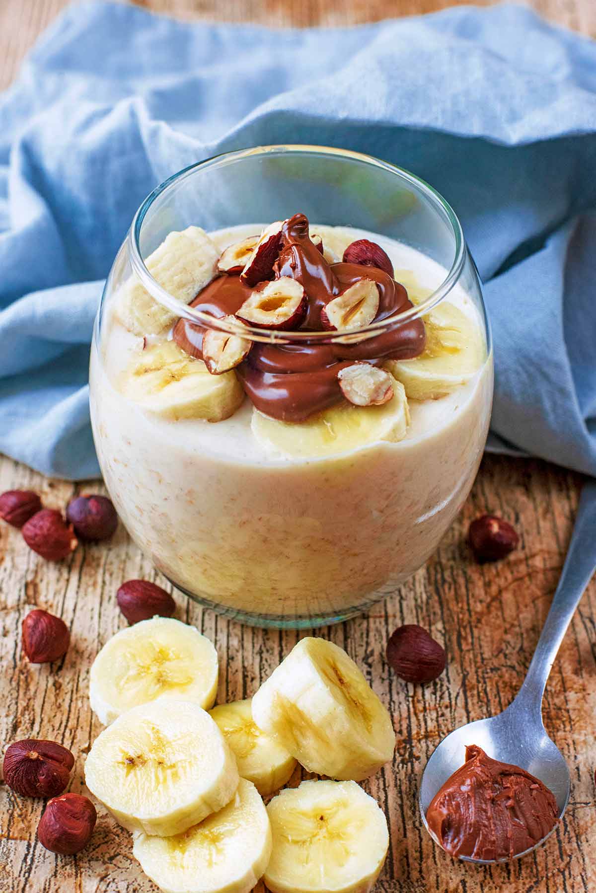 A glass of overnight oats topped with nutella, hazelnuts and banana slices.