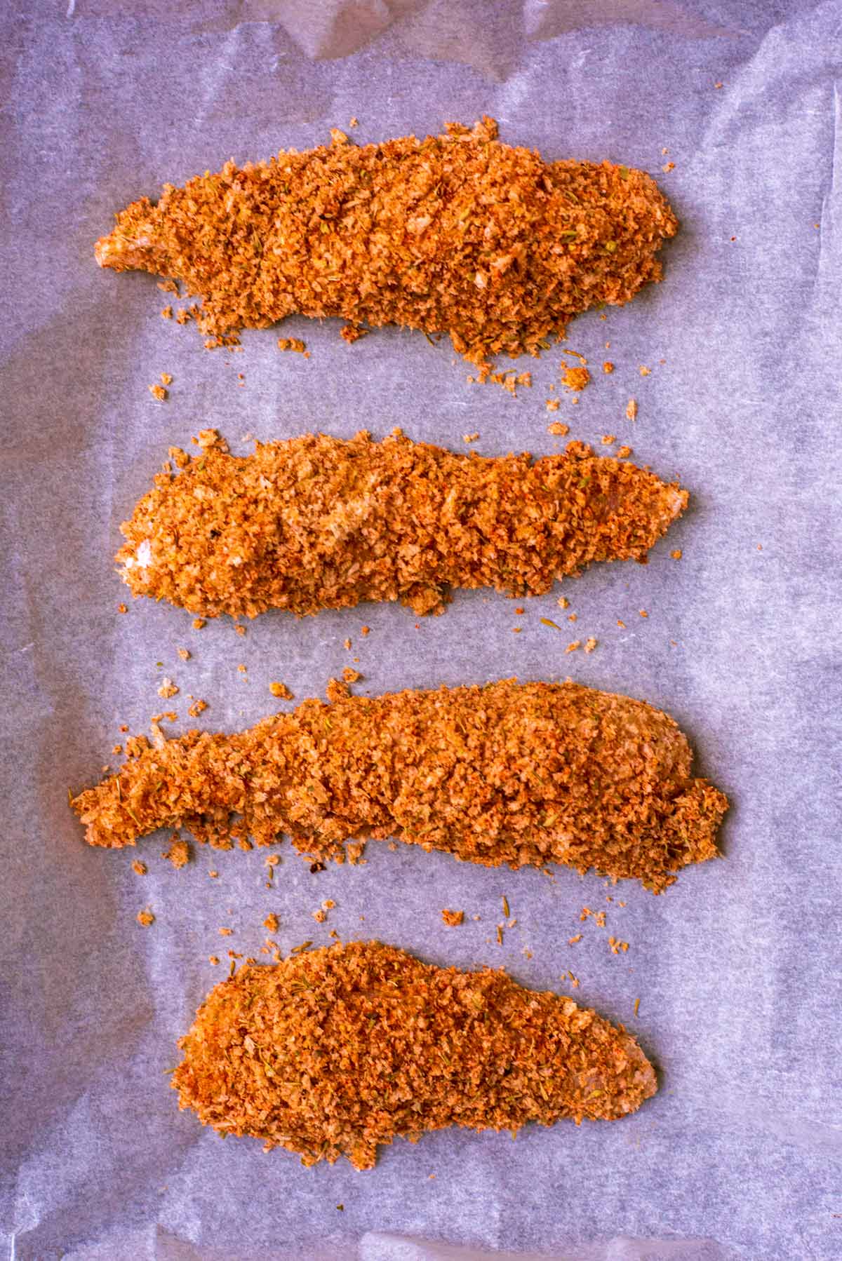Long chicken strips, coated in seasoned breadcrumbs, arranged in a row on a baking sheet.