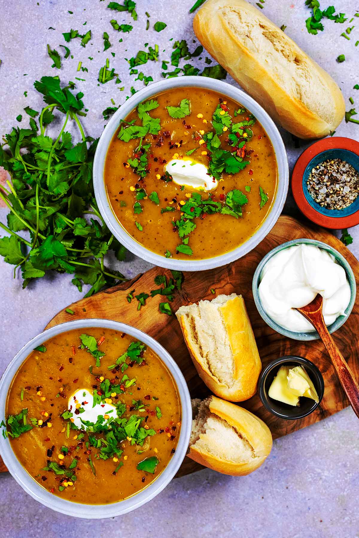 Two bowls of soup surrounded by bread and chopped herbs.