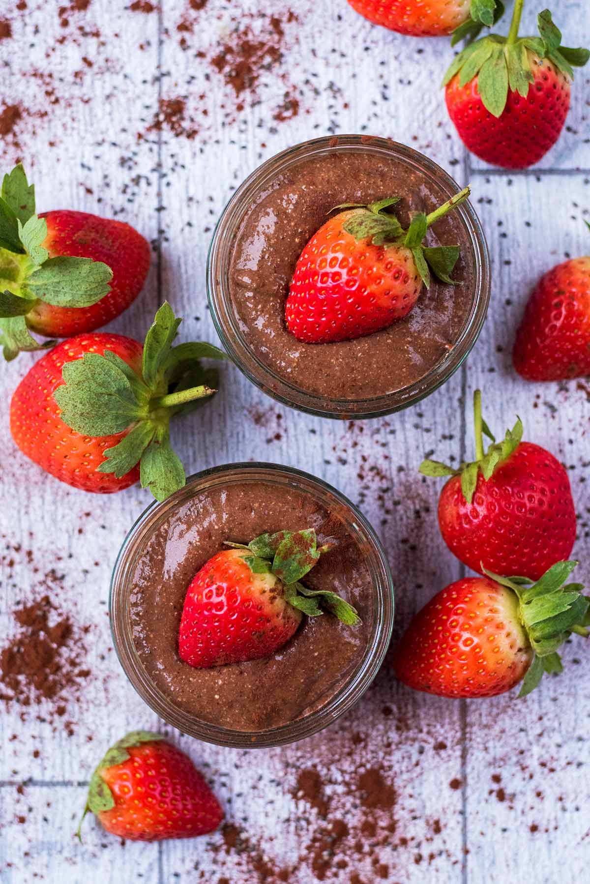 Chocolate pudding in two glass ramekins surrounded by strawberries and cocoa powder.