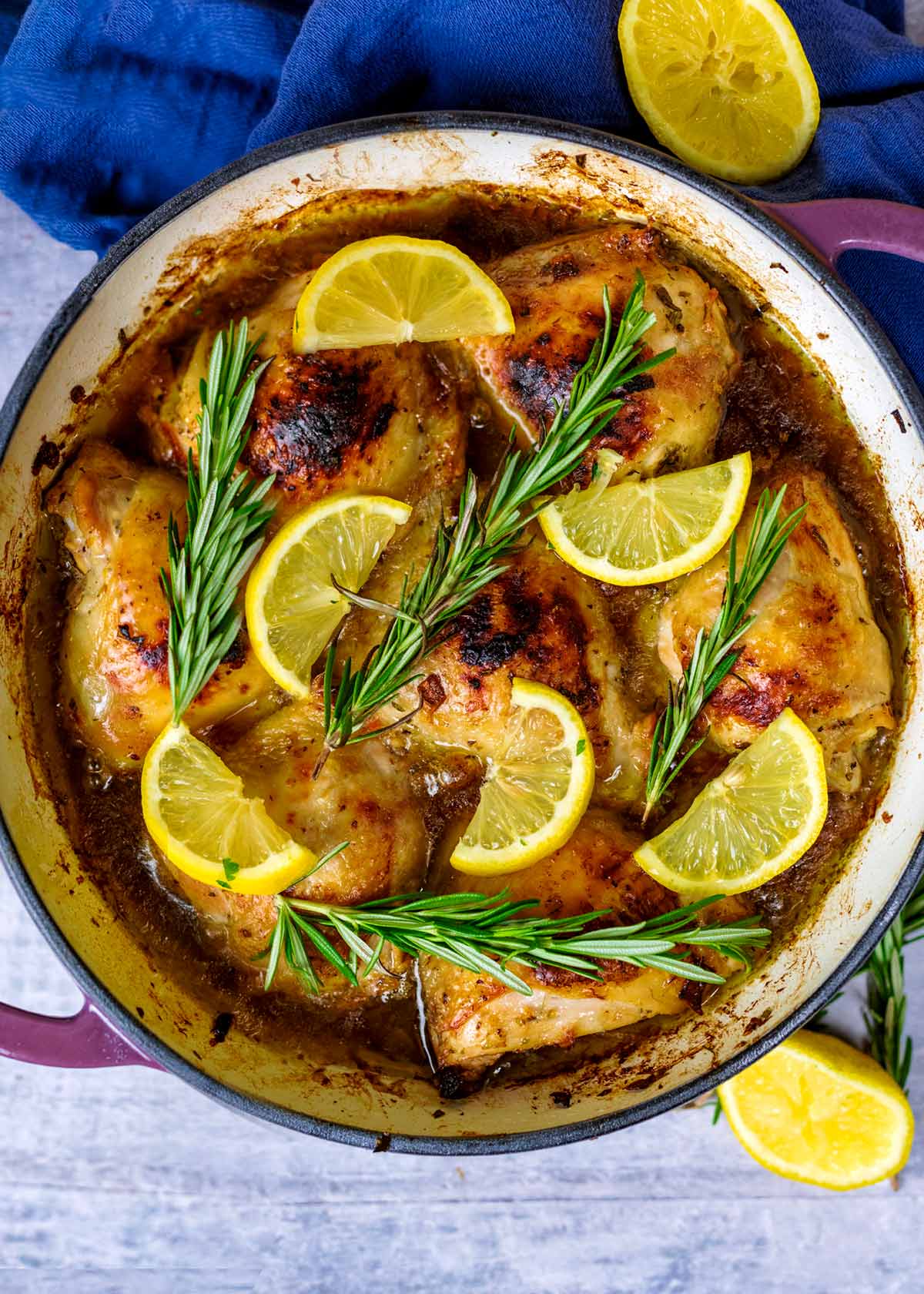 A large cooking pan containing cooked chicken thighs, lemon slices and rosemary sprigs.