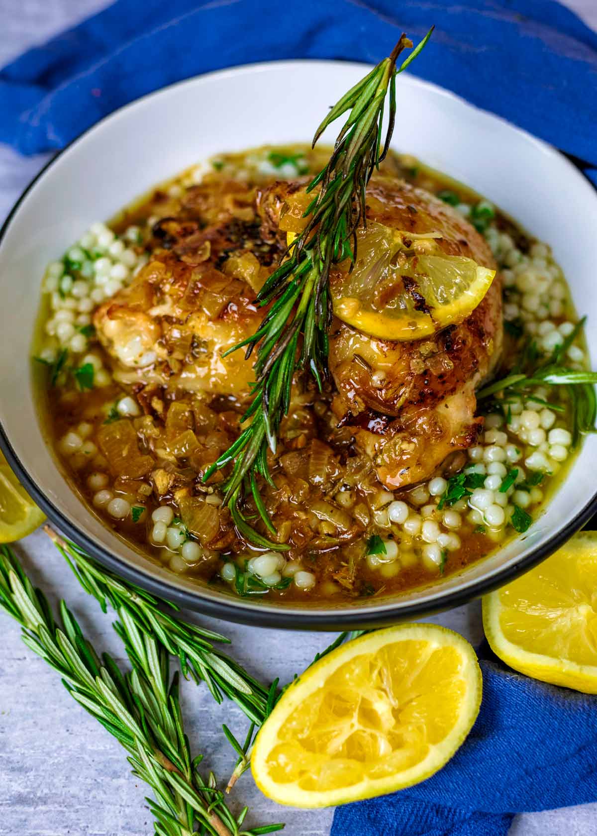 A cooked chicken thigh on top of couscous in a bowl with a sprig of rosemary on top.