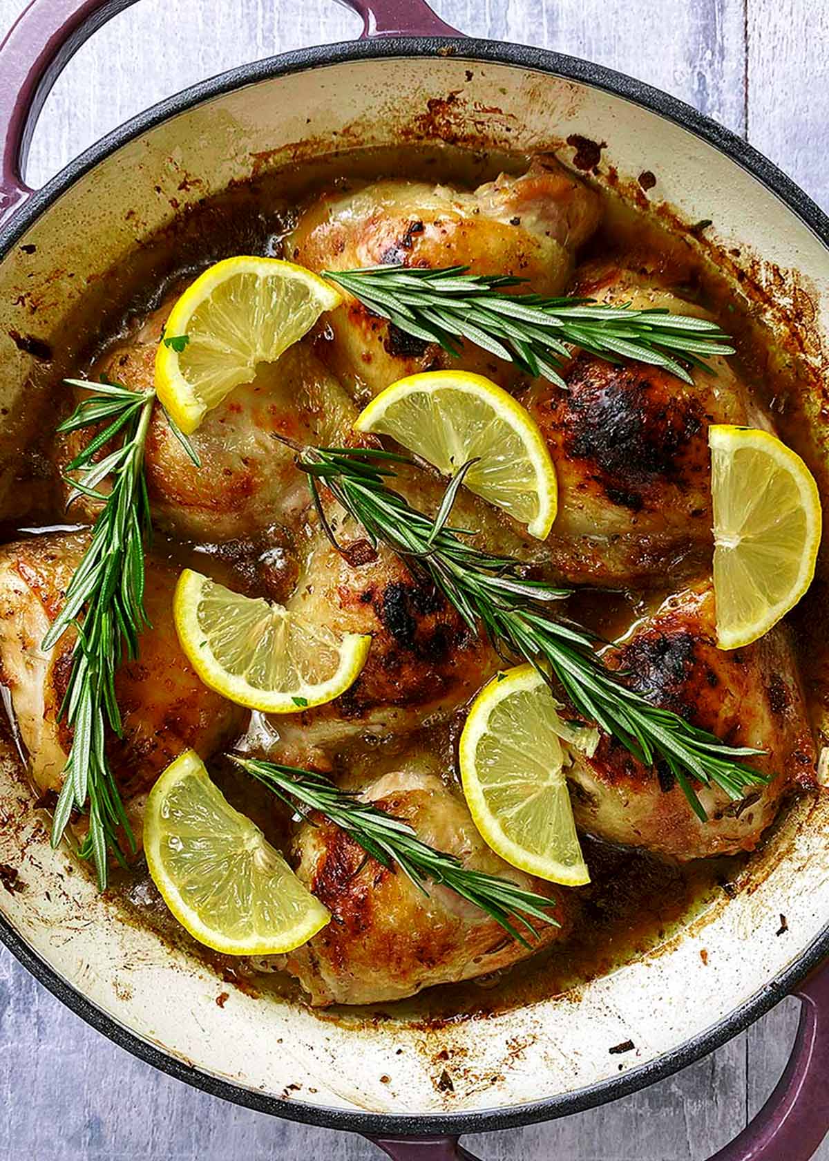 Cooked chicken thighs in a baking dish topped with lemon slices and sprigs of rosemary.