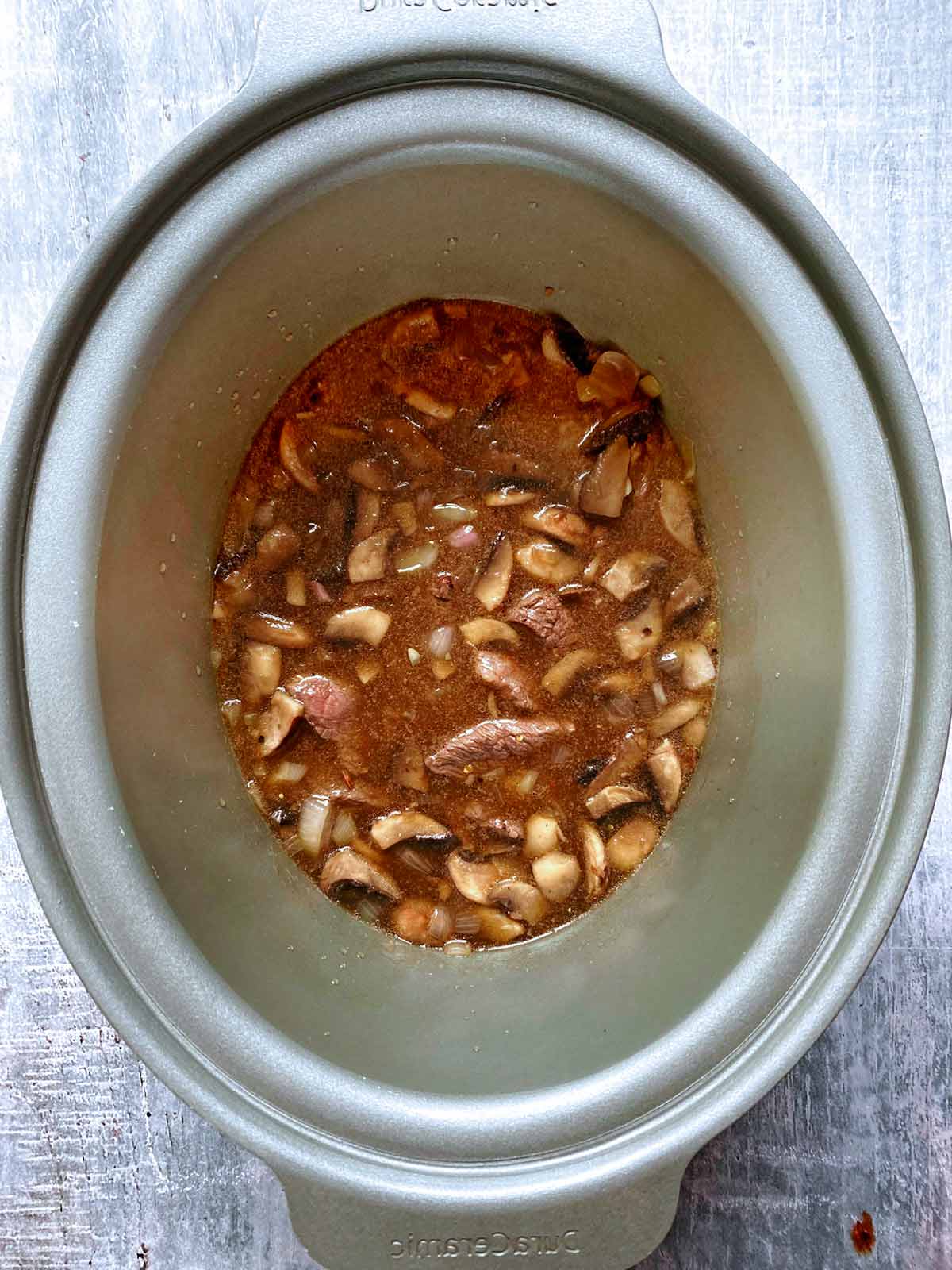 A slow cooker pot with cooked, beef, mushrooms and shallots all covered in stock.