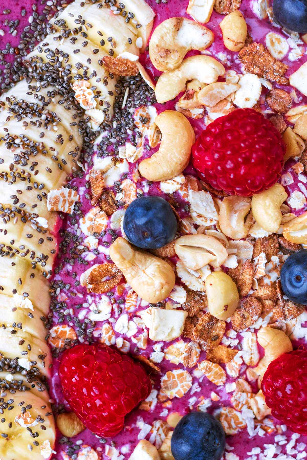 Raspberries, blueberries, a sliced banana, nuts and seeds topping a purple smoothie.