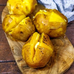 Four slow cooker jacket potatoes on a wooden board with butter melting in them.