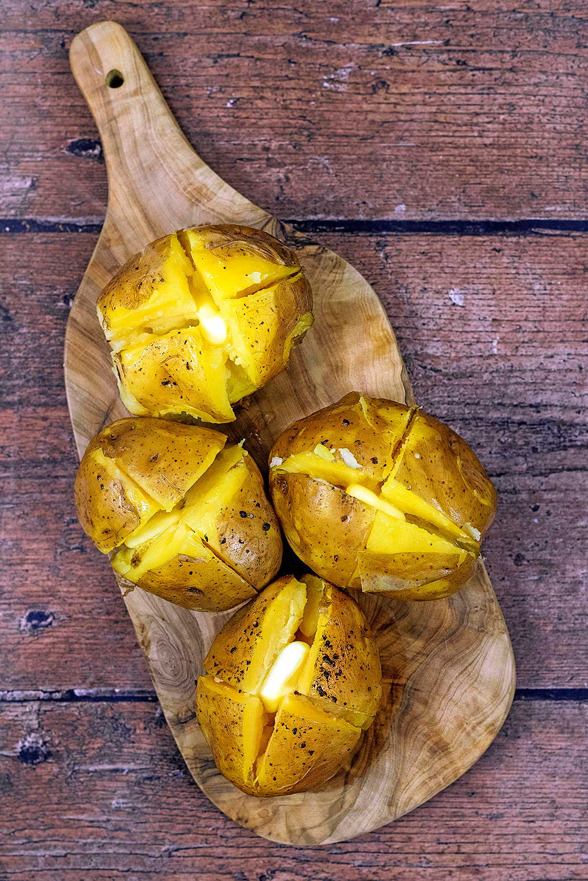 Four baked potatoes on a board, cut open with melted butter in them.