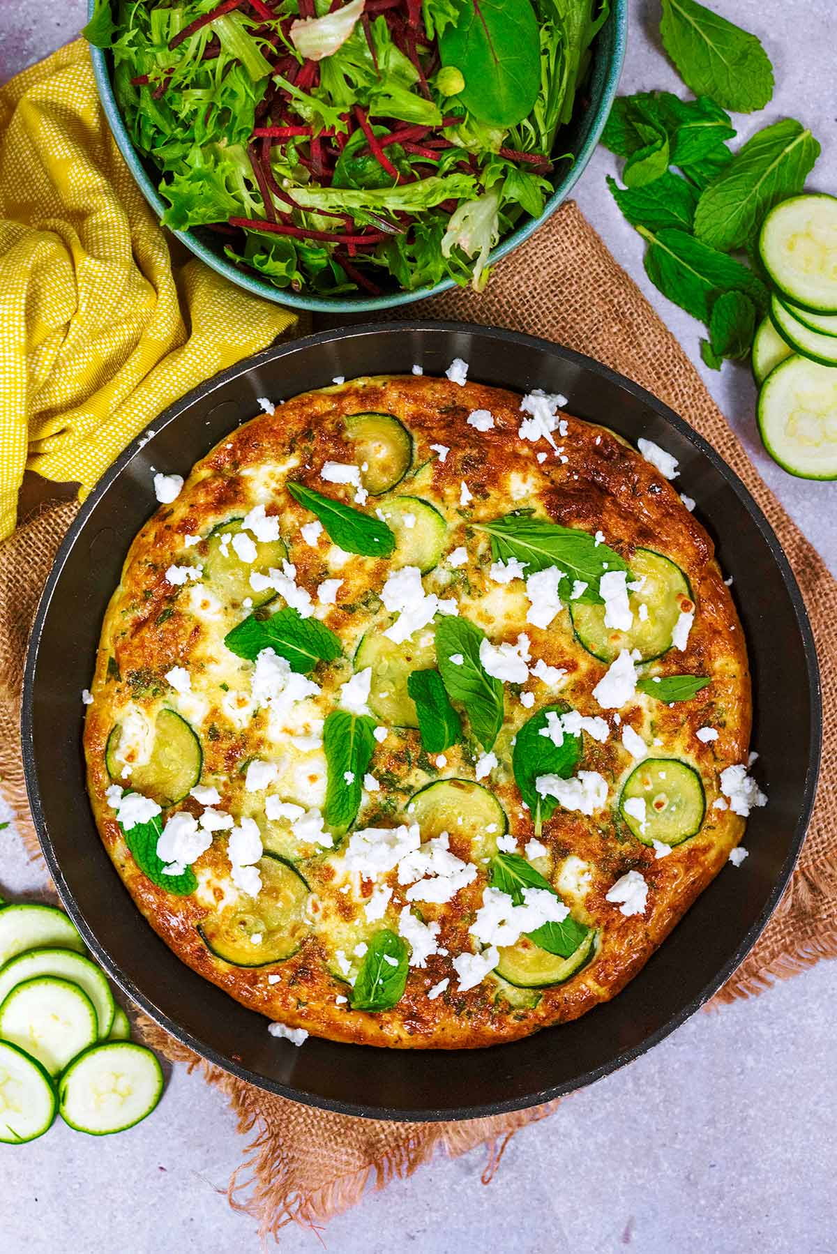 A frittata in a pan with a yellow towel wrapped around the handle. A bowl of salad is next to it.