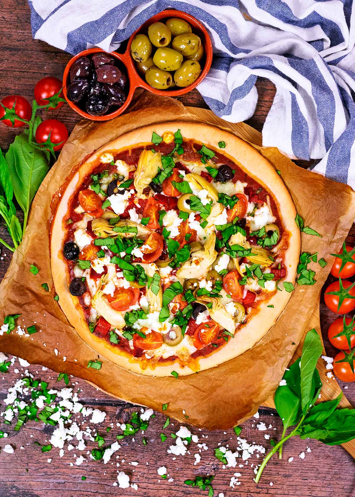 A whole pizza on a square of baking paper surrounded by olives, tomatoes and basil.
