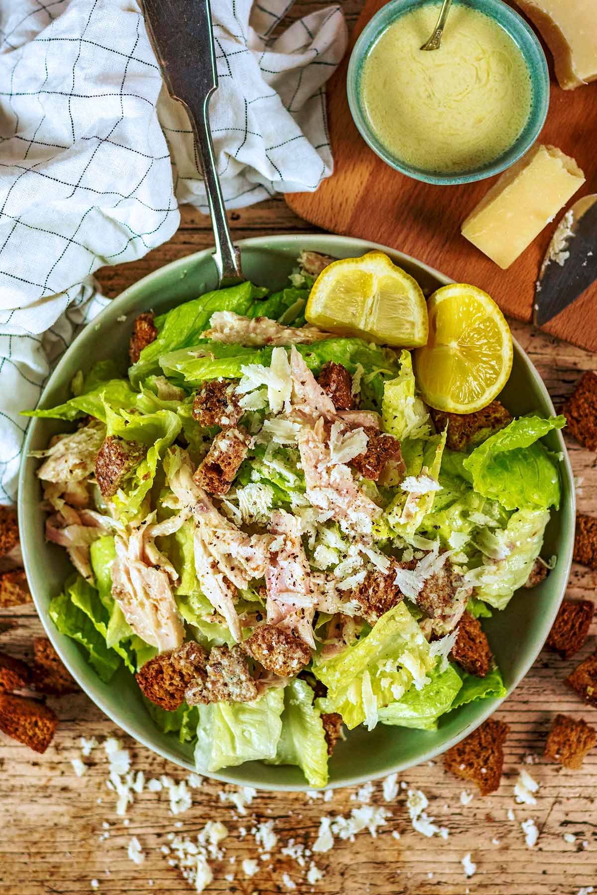 A bowl of caesar salad next to a board with cheese, a knife and a bowl of caesar dressing.