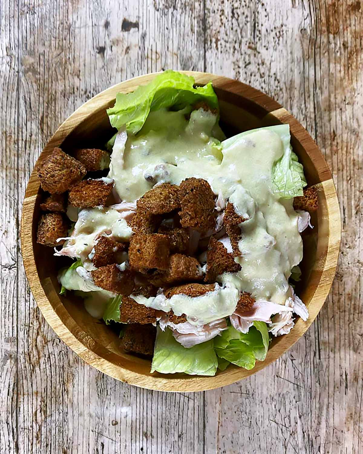 Chopped lettuce, croutons and chopped chicken in a bowl covered in caesar dressing.