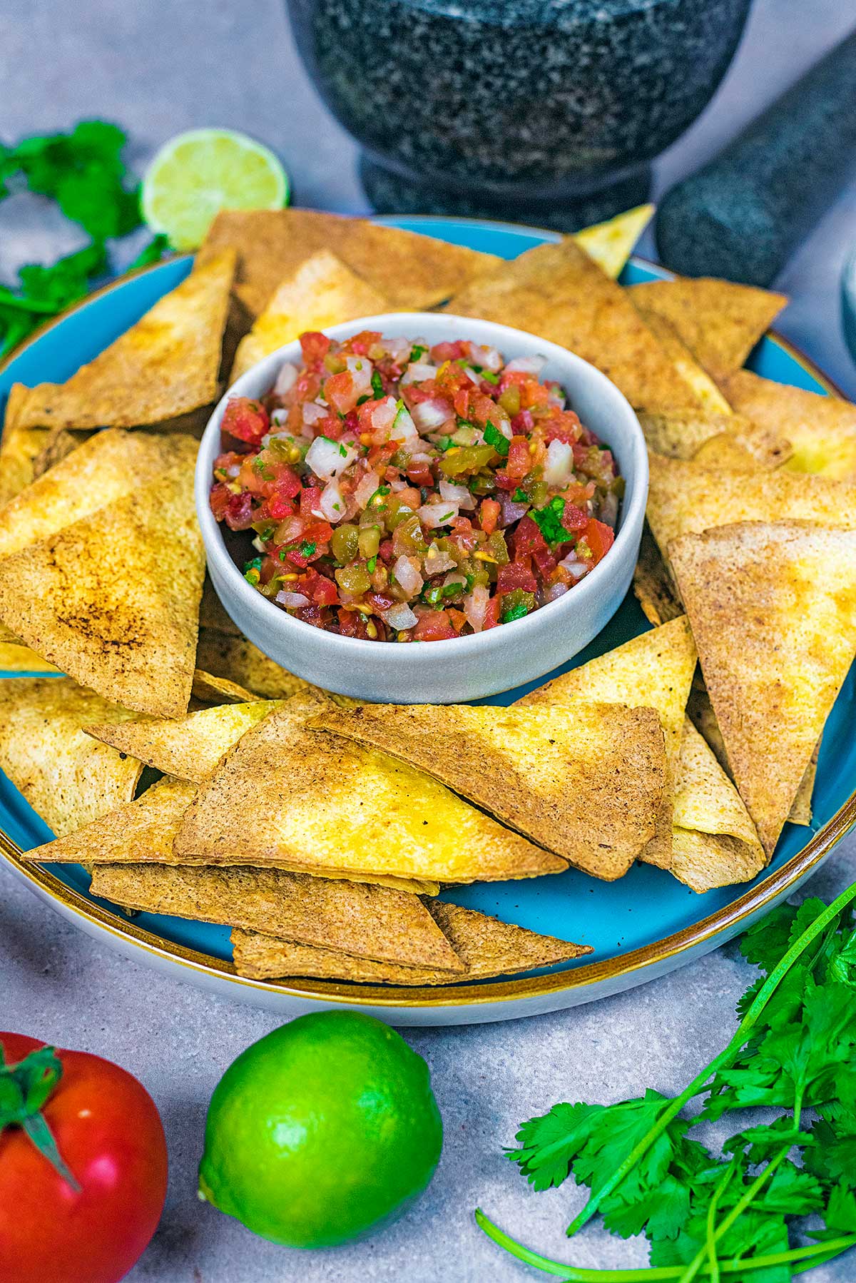 A plate of tortilla chips and salsa next to some limes and tomatoes.