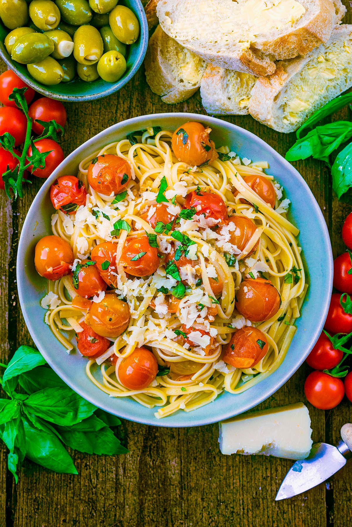 A bowl of tomato linguine next to a bowl of olives, some bread and some basil leaves.