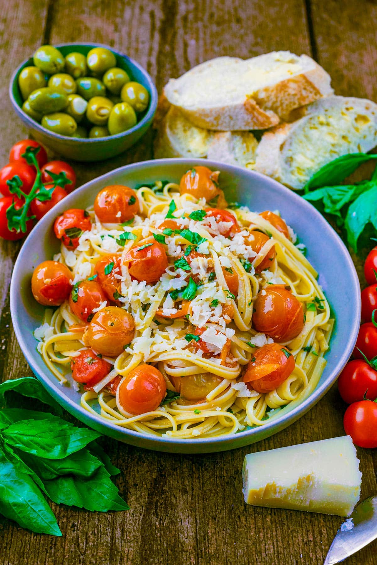 A bowl of pasta and tomatoes in front of a bowl of olives and some slices of buttered bread.