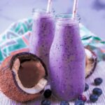 Two small glass bottles full of blueberry and coconut smoothie next to a broken open coconut