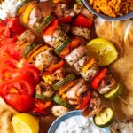 Greek chicken skewers on a wooden board surrounded by side dishes.