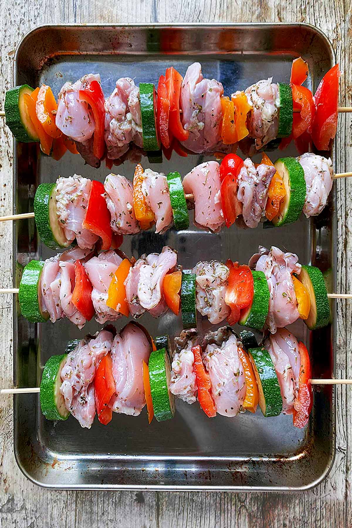 A metal baking tray with four chicken and vegetable skewers on it.