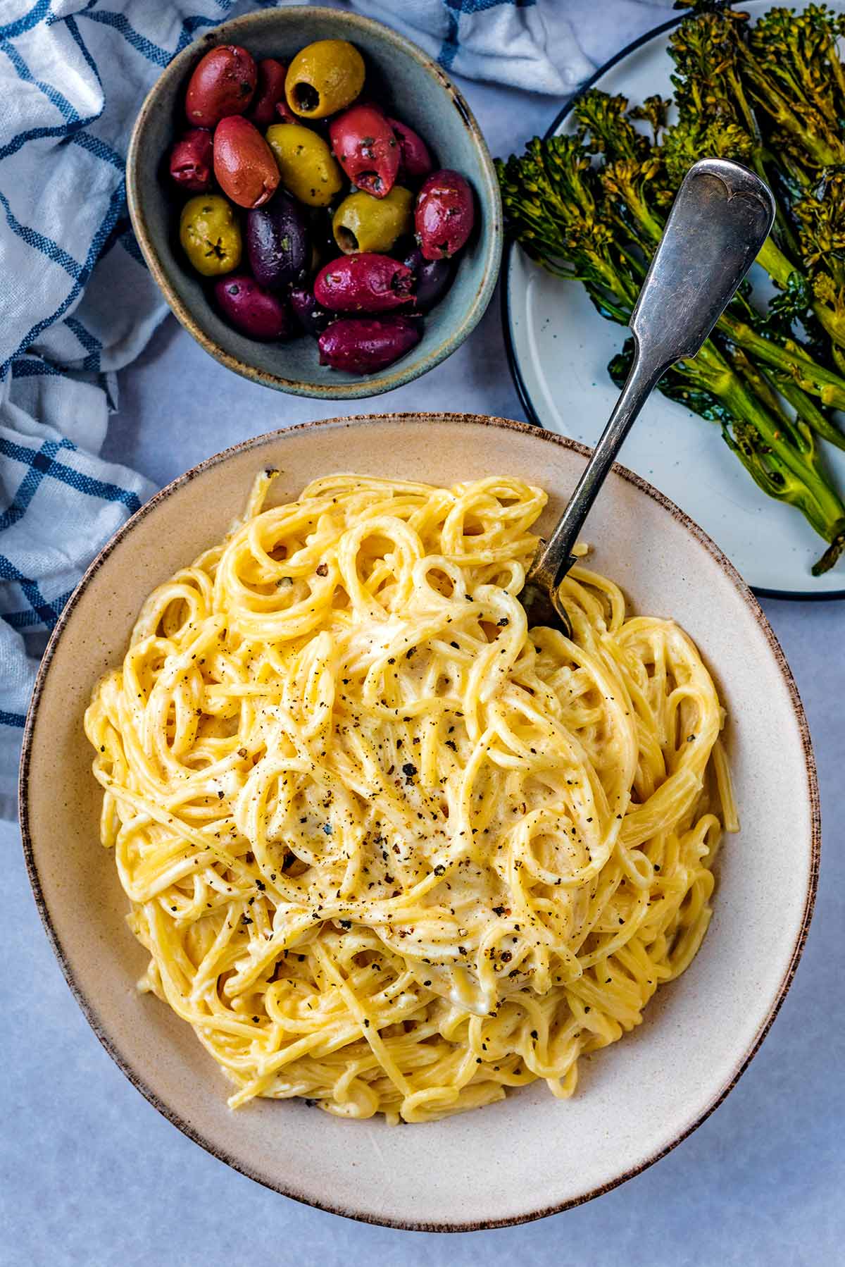 A large bowl of creamy spaghetti next to a bowl of olives and some broccoli.