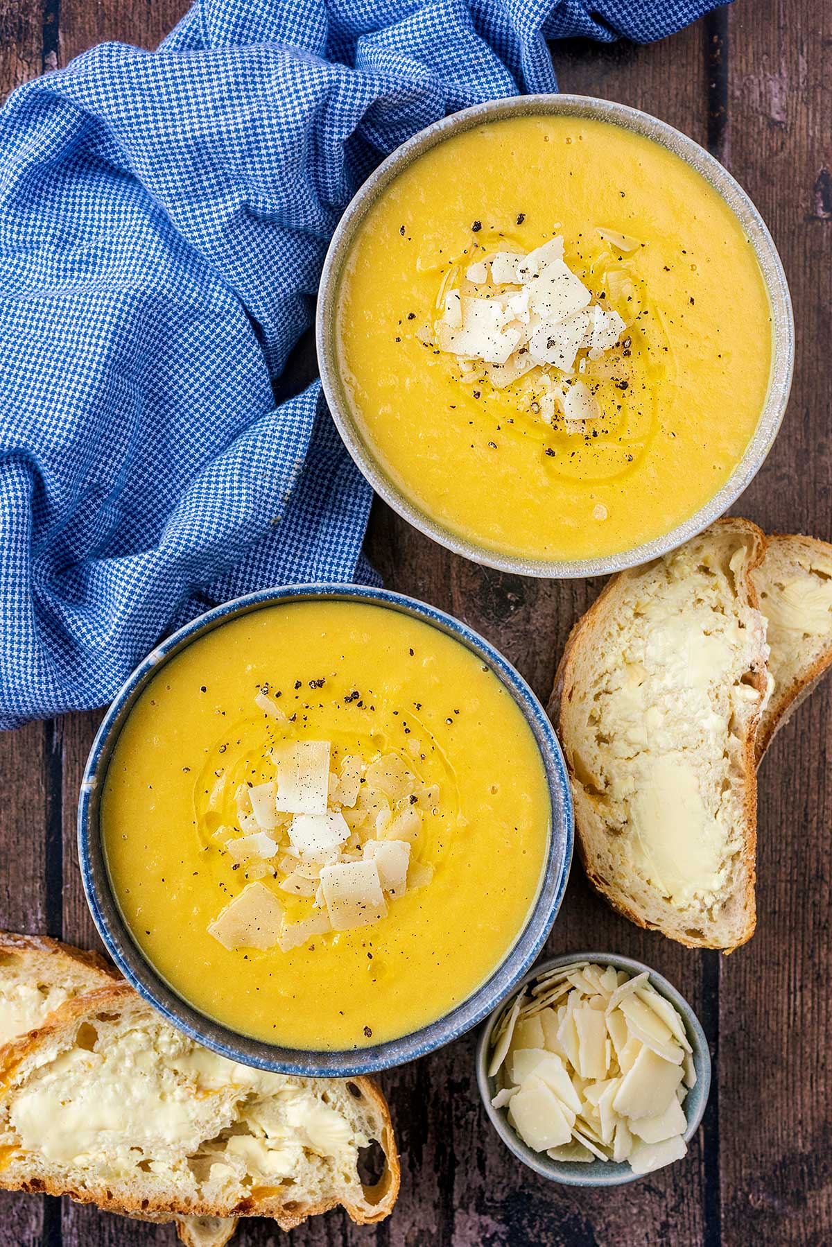 Two bowls of yellow coloured soup next to buttered bread and a small pot of Parmesan shavings.