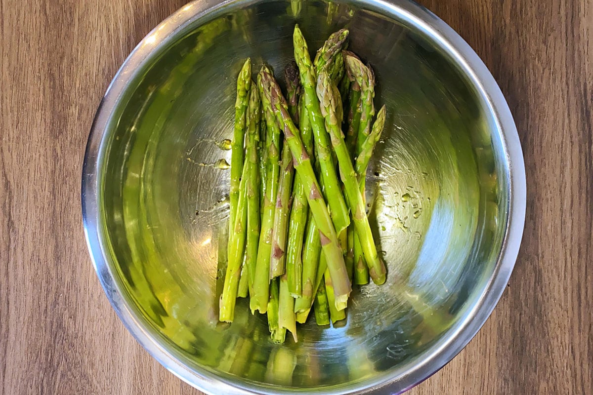 A metal mixing bowl containing asparagus spears coated in oil.