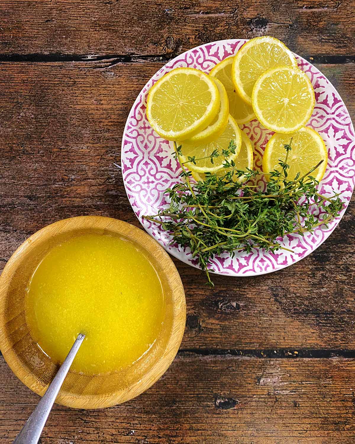 A small bowl of melted butter next to a plate of lemon slices and sprigs of thyme.
