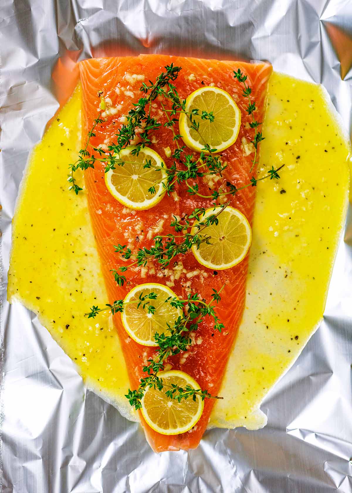 A whole side of salmon on a sheet of foil covered in a buttery sauce, lemon slices and thyme sprigs.