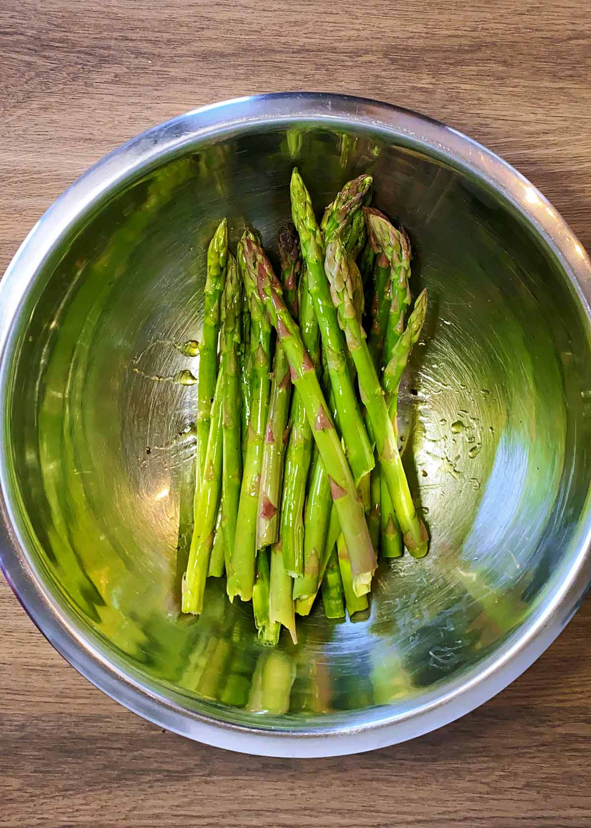 A metal mixing bowl containing asparagus spears coated in oil.