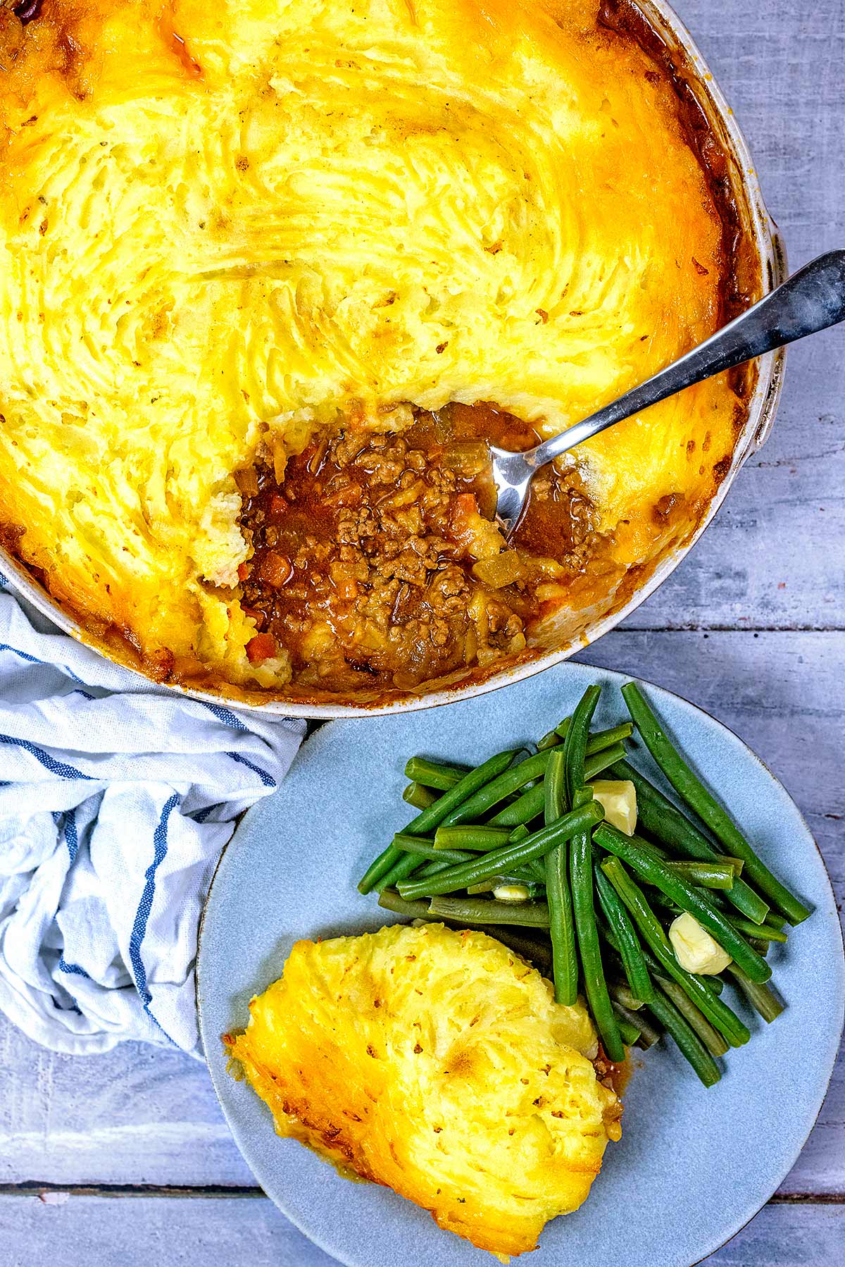 A large serving dish of shepherd's pie next to a plate of more pie and some green beans.