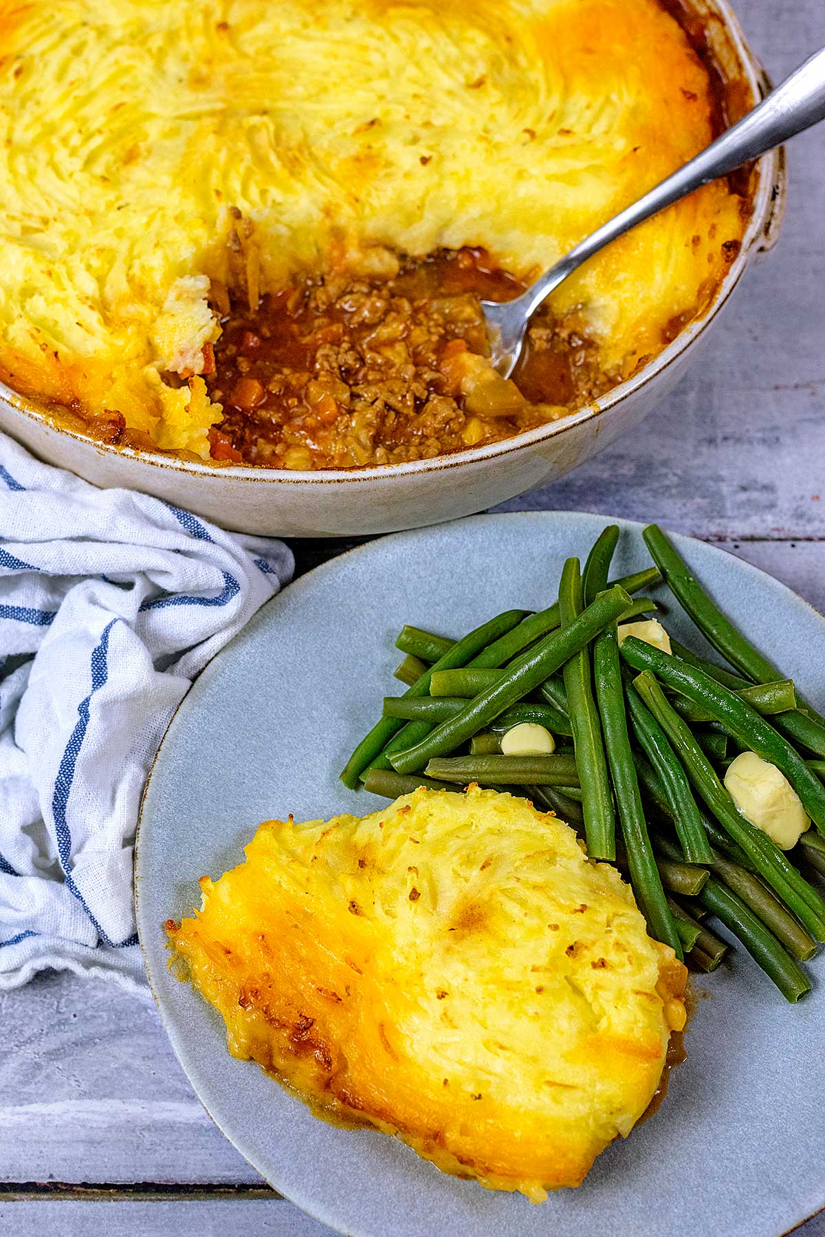 A plate of shepherd's pie and green beans in front of a large dish of more pie.