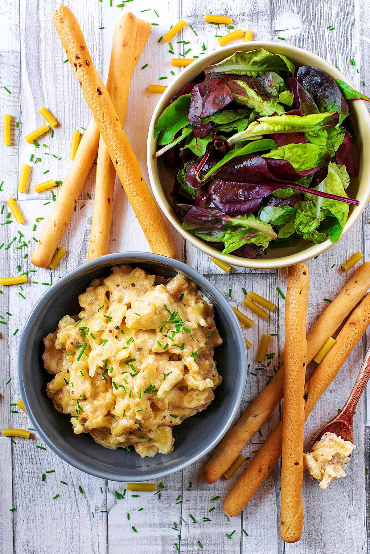 A bowl of mac and cheese next to a bowl of salad leaves and some bread sticks.