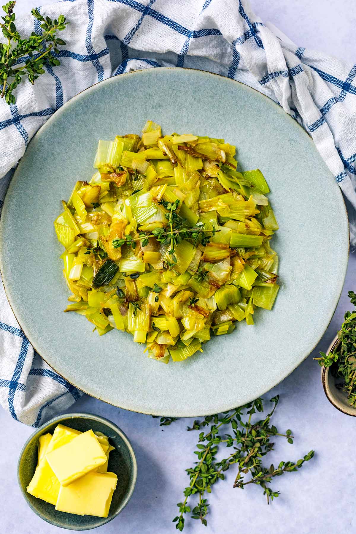 A plate of cooked leeks next to a bowl of butter and a bowl of fresh thyme sprigs.