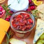 Christmas Chutney in a jar. Two cheese on crackers sit in front and grapes in the background.