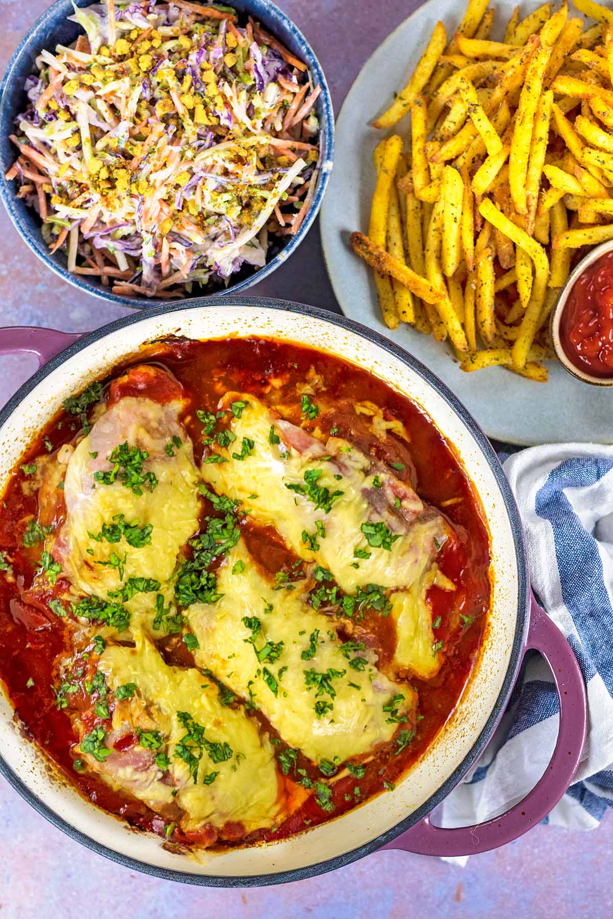 A baking dish containing hunter's chicken next to a plate of fries and bowl of salad.