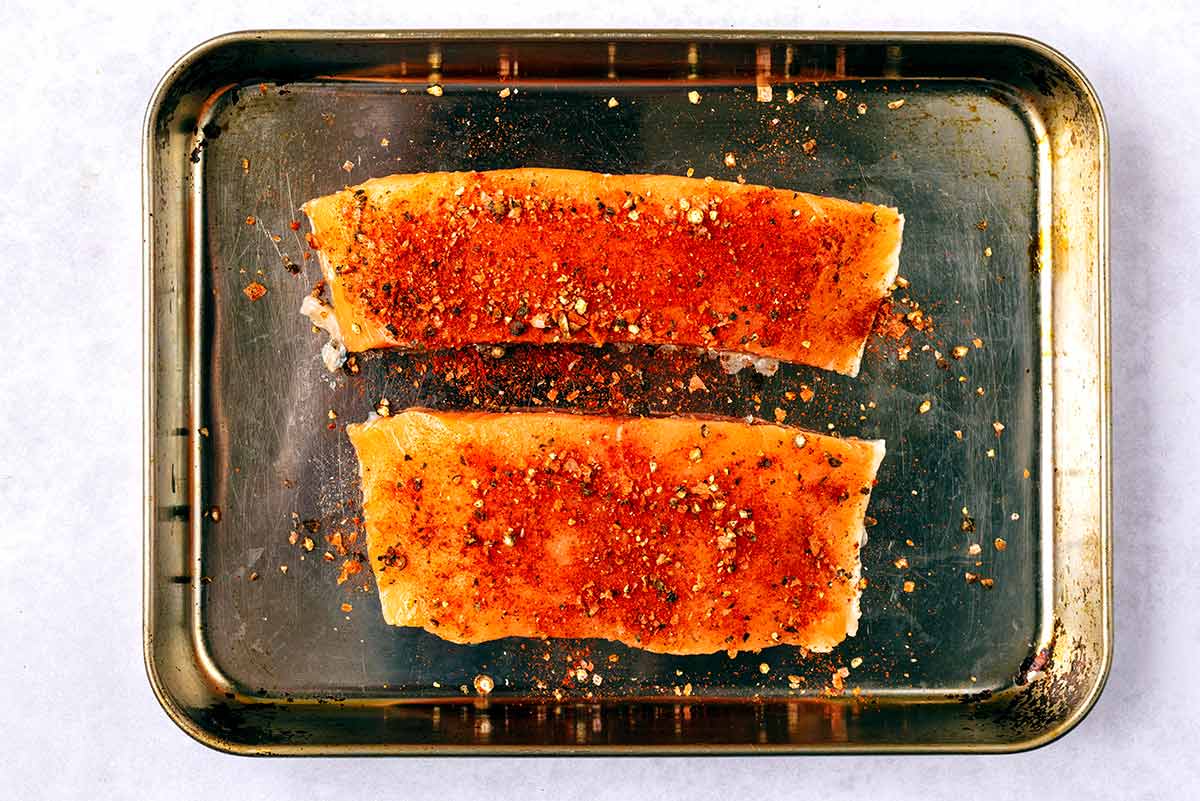 Two salmon fillets covered in seasoning on a baking tray.