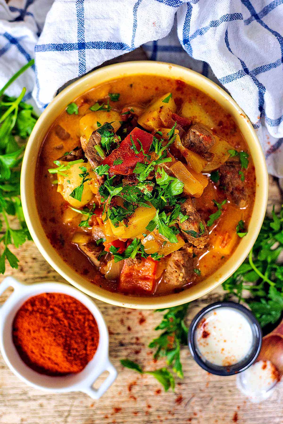 A bowl of beef goulash next to a small pot of cream and a dish of paprika.