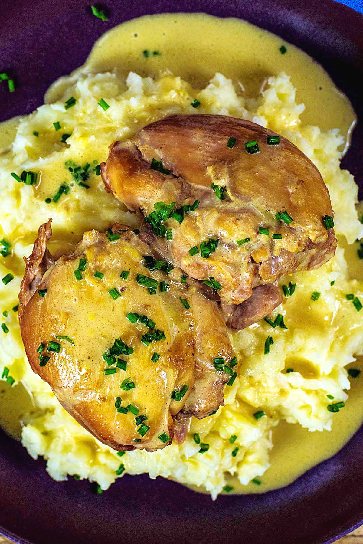 Two cooked chicken thighs on a bed of mashed potato covered in a creamy sauce with chopped chives.