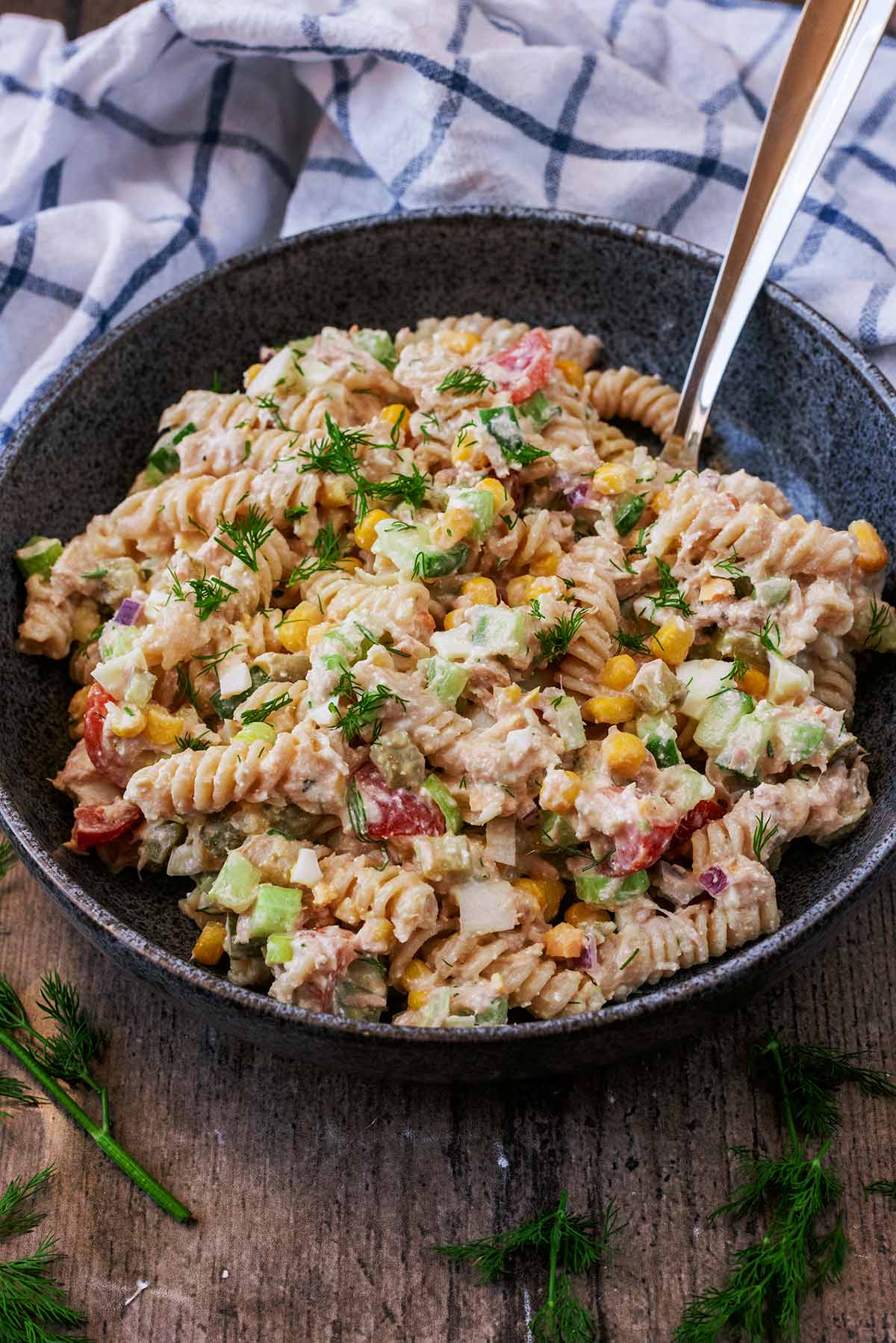 Pasta salad in a bowl with a spoon in front of a checked towel.