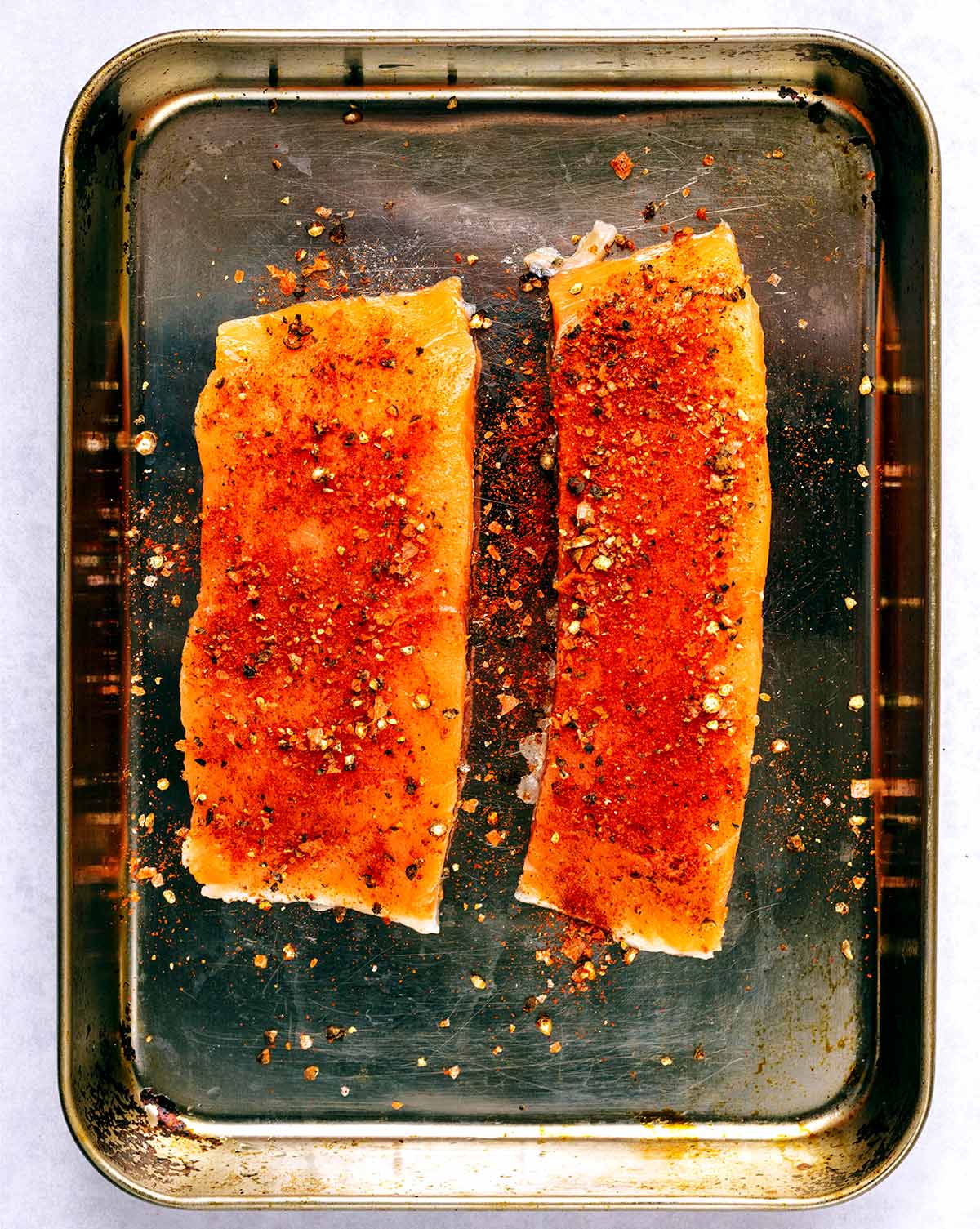 Two salmon fillets covered in seasoning on a baking tray.