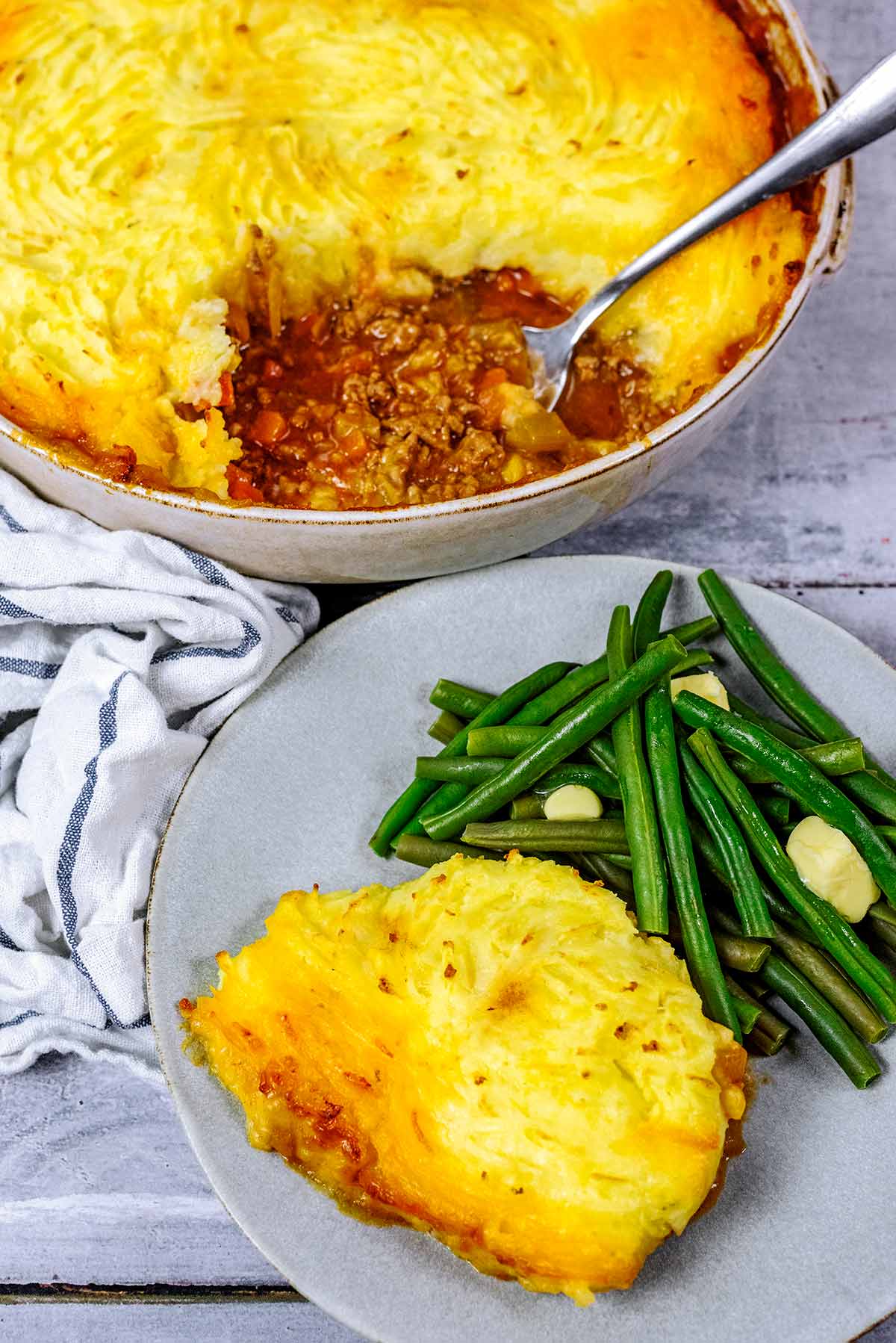 A plate of shepherd's pie and green beans in front of a large dish of more pie.