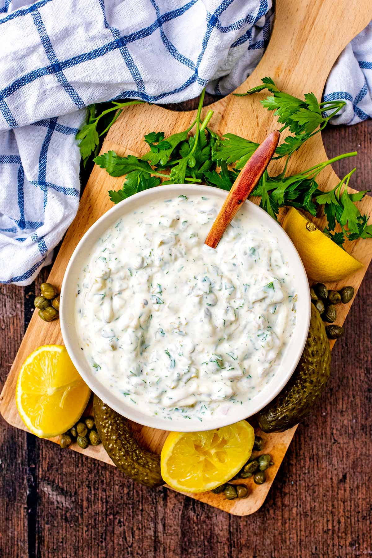 A bowl of tartare sauce on a board with parsley, lemons, gherkins and capers.