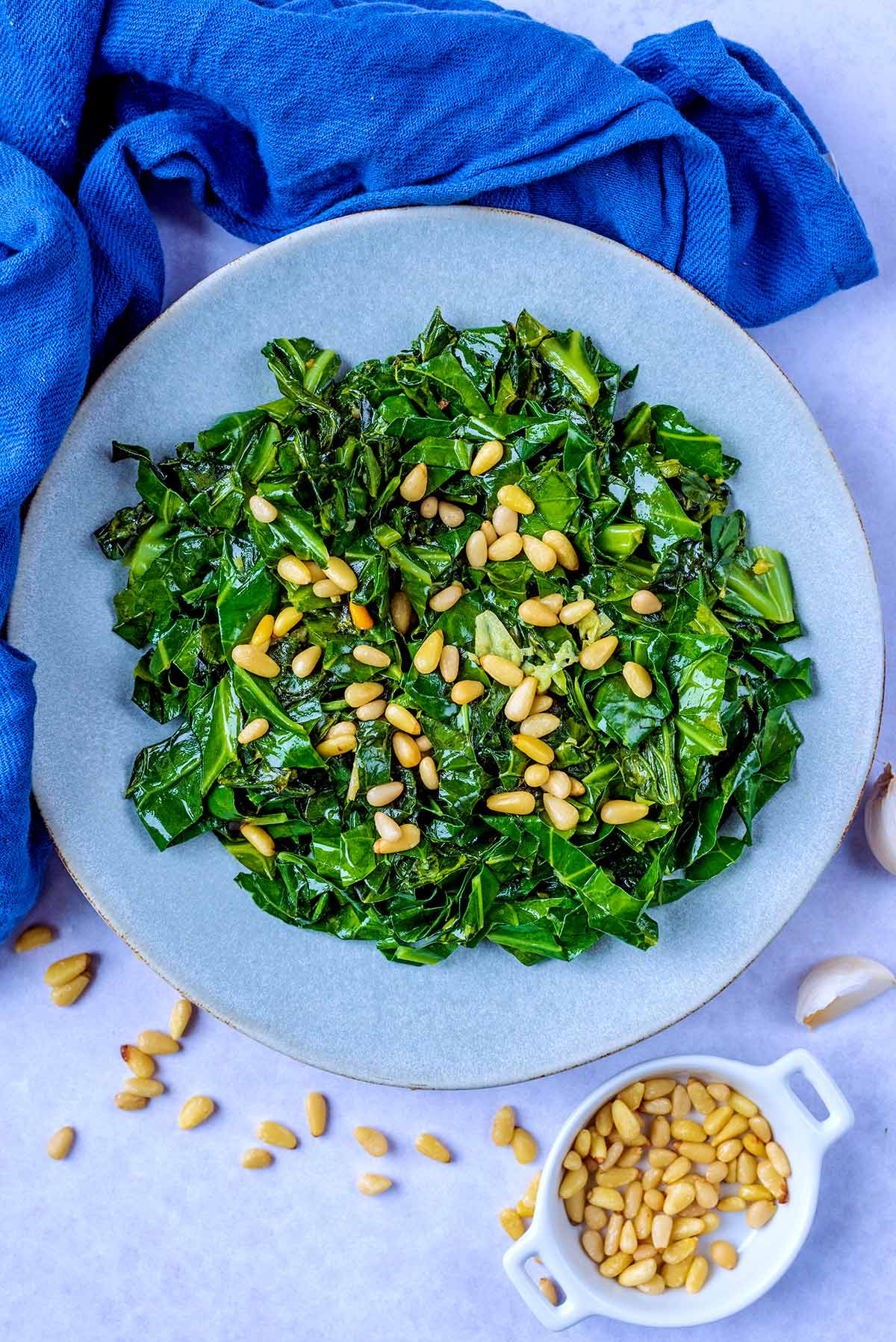 A plate of cooked spring greens next to a blue towel and a small dish of pine nuts.