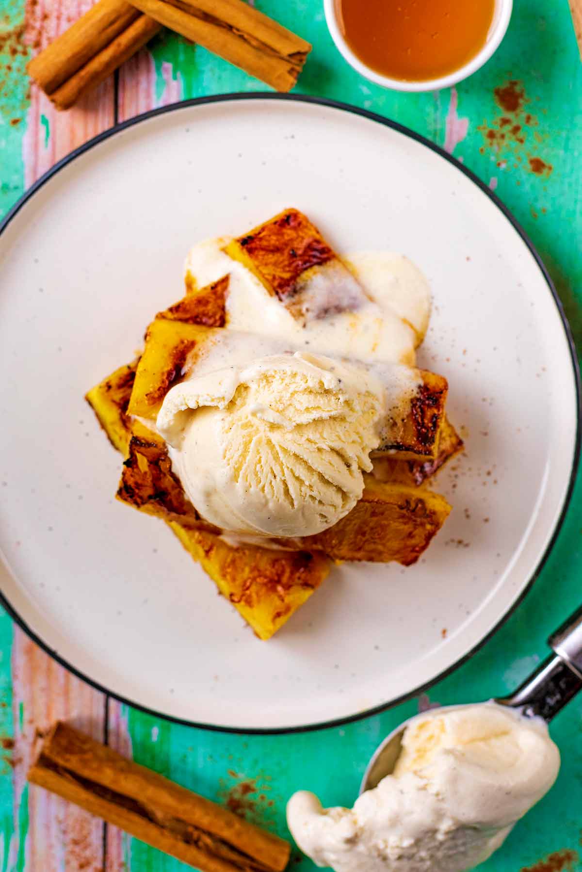 A plate with grilled pineapple and ice cream surrounded by cinnamon sticks and a pot of honey.