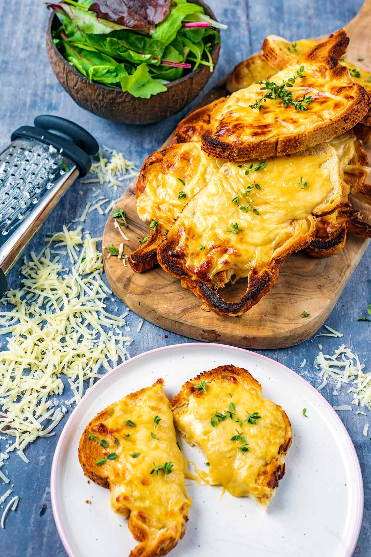 Welsh rarebit on a wooden serving board next to a cheese grater and a bowl of salad.