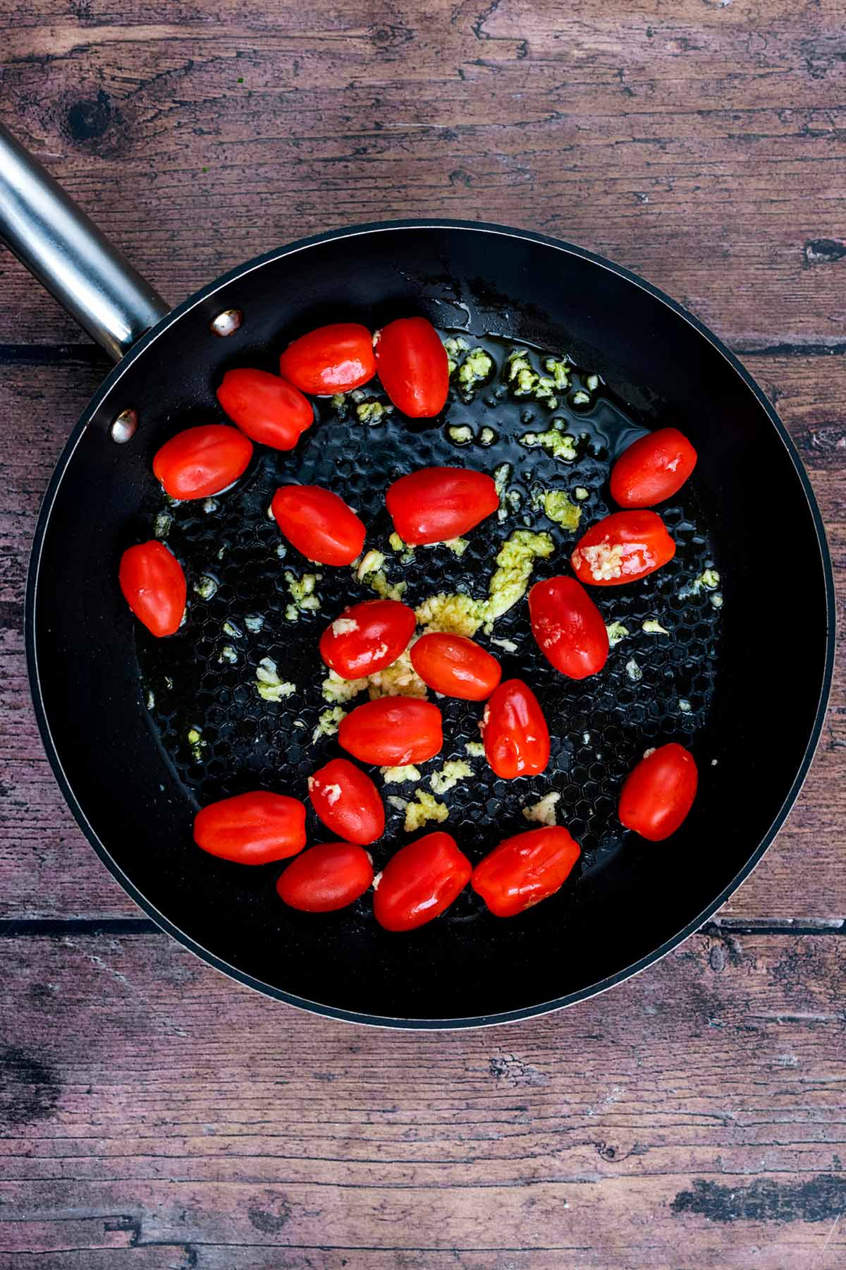 Cherry tomatoes and crushed garlic cooking in a black frying pan.