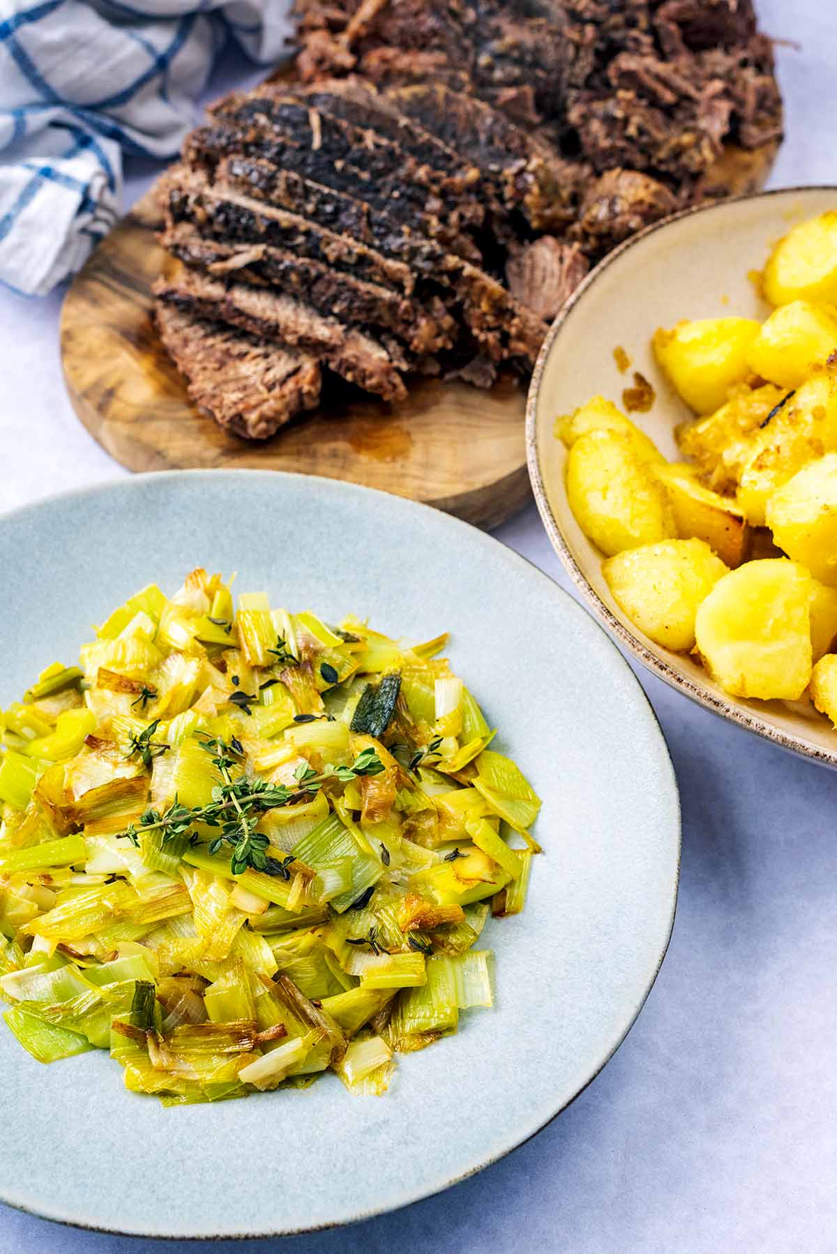 A plate of cooked leeks in front of a bowl of roast potatoes and some sliced beef on a board.