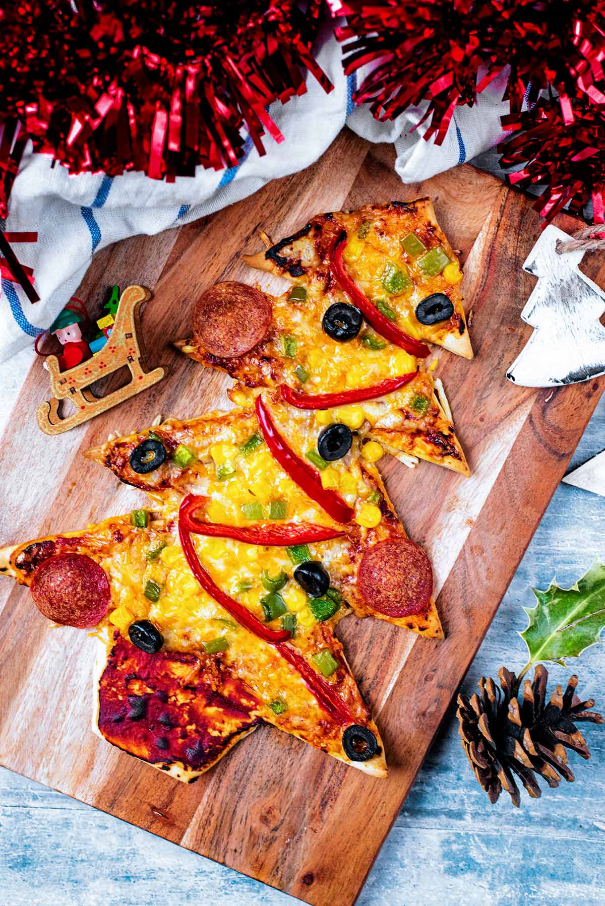 A Christmas Tree shaped Pizza on a wooden board with red tinsel next to it.