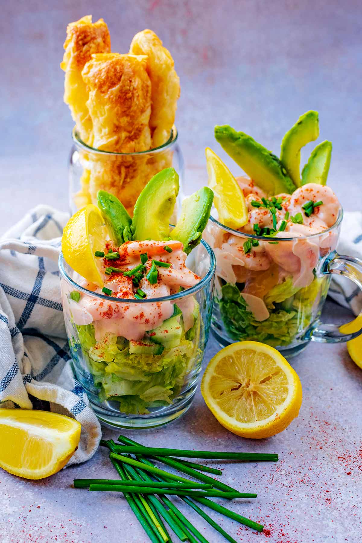 Two glasses containing prawn cocktails surrounded by lemons and chives.