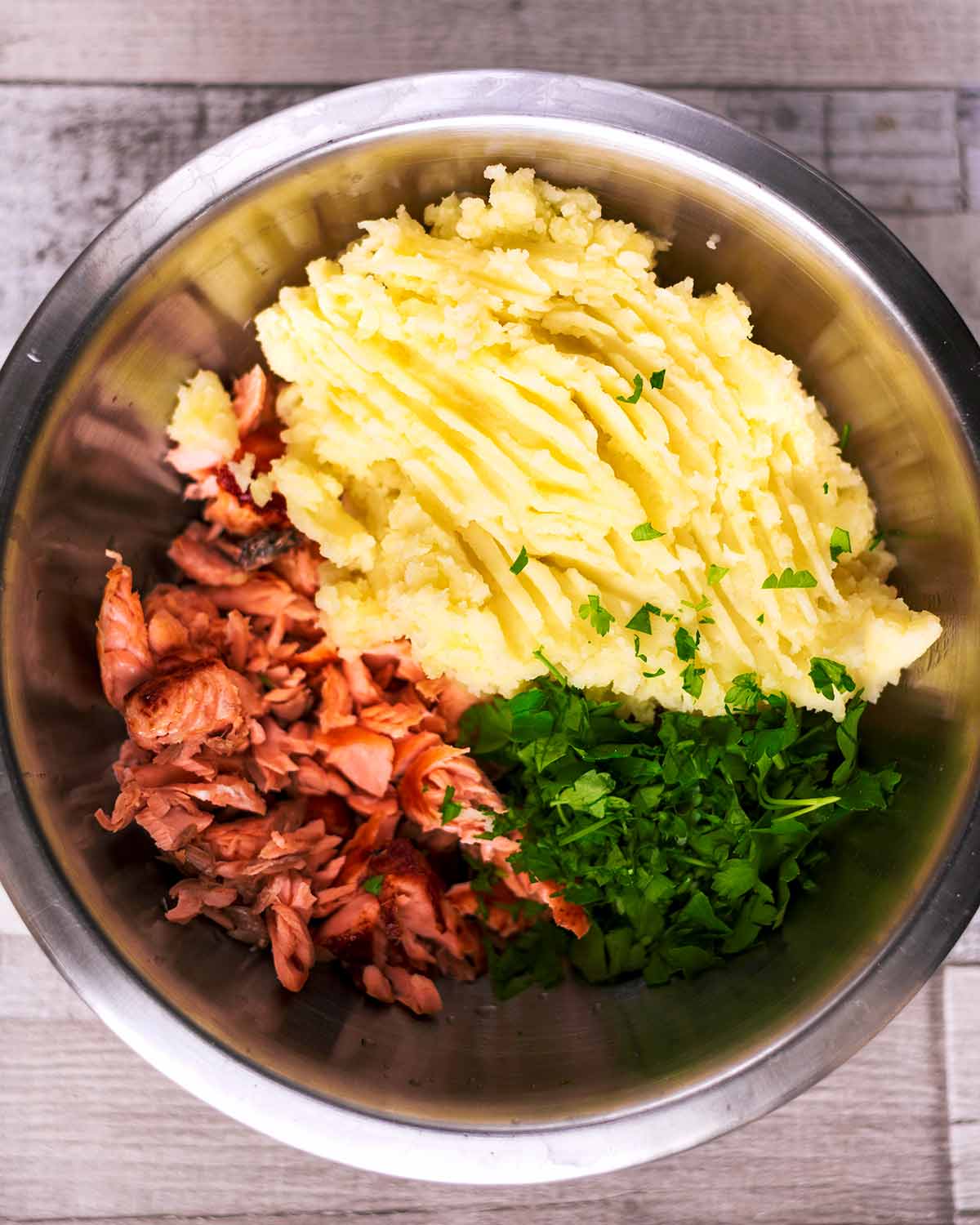 Flaked salmon, mashed potato and chopped herbs in a large mixing bowl.